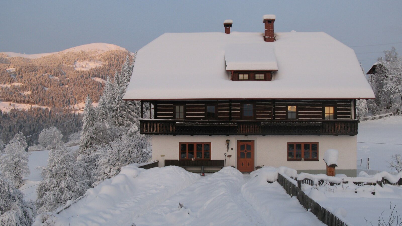 Der verschneite Bauernhof mit Holzbalkon und leuchtenden Fenstern, umgeben von schneebedeckten Bergen und Wäldern.