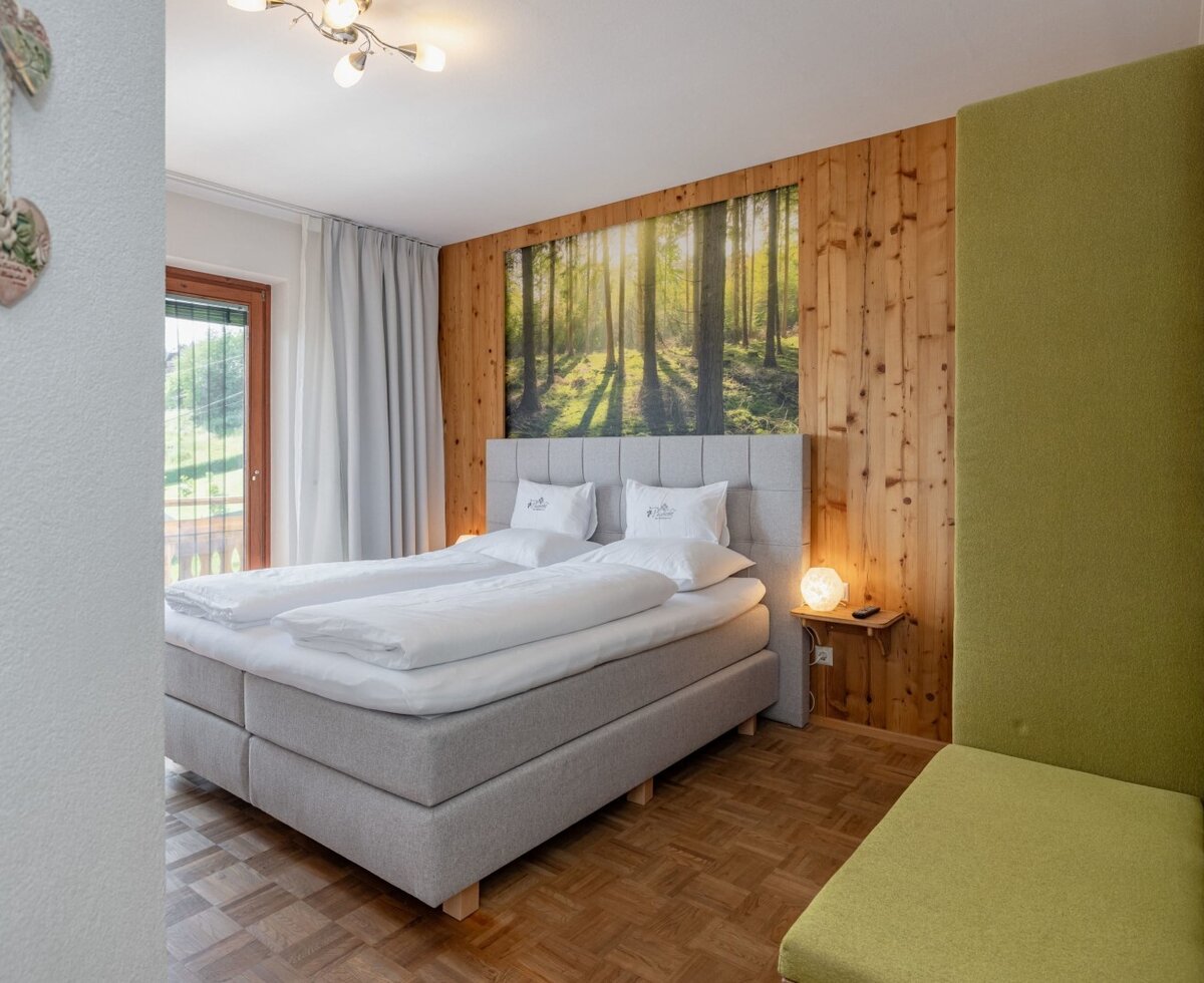 Doppelbett im Schlafzimmer des Bauernhofs mit Holzwand, einem Wandbild mit Waldmotiv, einer gepolsterten Sitzbank und Zugang zum Balkon.