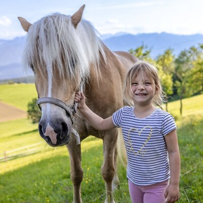 Ein Kind streichelt auf dem Bauernhof ein Pferd, umgeben von grünen Feldern und Bergen.