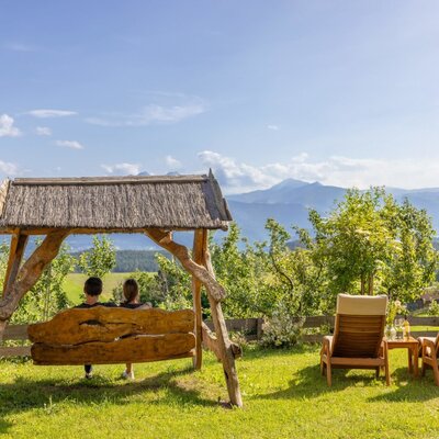 Sitzbereich im Freien auf dem Bauernhof mit einer Holzschaukel, zwei Liegestühlen und Bergblick.