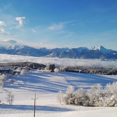 Der Bauernhof liegt in einer winterlichen Landschaft mit schneebedeckten Feldern, Bergen und Talnebel.