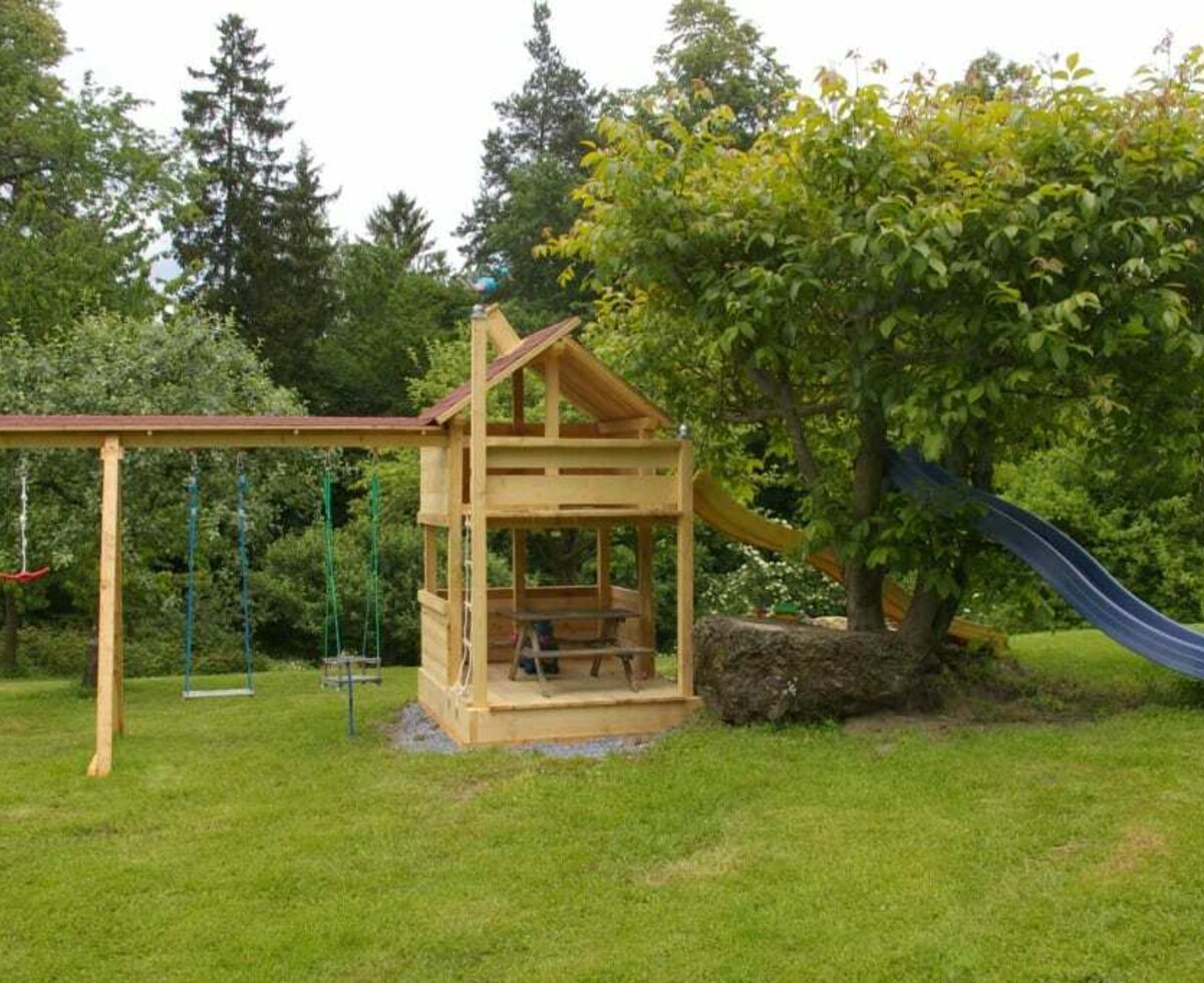 Der Bauernhof bietet einen Kinderspielplatz mit Schaukeln, Rutschen und einem Spielhaus auf einer Rasenfläche.