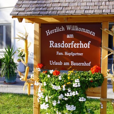 Das Willkommensschild des Bauernhofs Rasdorferhof, dekoriert mit Blumen und Maiskolben, vor dem Gebäude mit Außenterrasse.