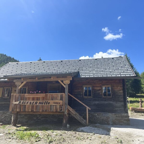 Eine urige Alm auf 1666 Metern Höhe mit einer gemütlichen Holzterrasse unter strahlend blauem Himmel.