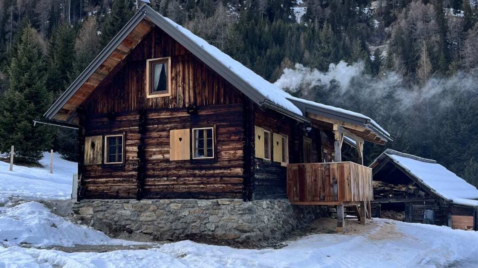 Eine urige Alm in einer verschneiten Berglandschaft mit einem gemütlichen Balkon vor einem bewaldeten Berghang.