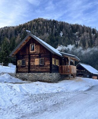 Eine urige Alm in einer verschneiten Berglandschaft mit einem gemütlichen Balkon vor einem bewaldeten Berghang.