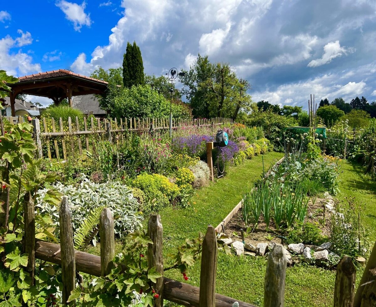 Der Garten des Bauernhofs mit Holzzaun, vielfältigen Blumenbeeten und einem Gemüseanbau.