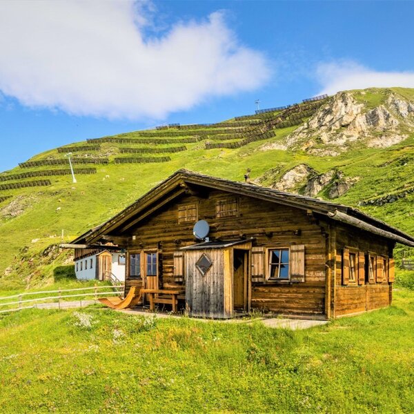 Die Almhütte in Holzbauweise liegt auf einem grünen Berghang und bietet einen Sitzbereich im Freien.
