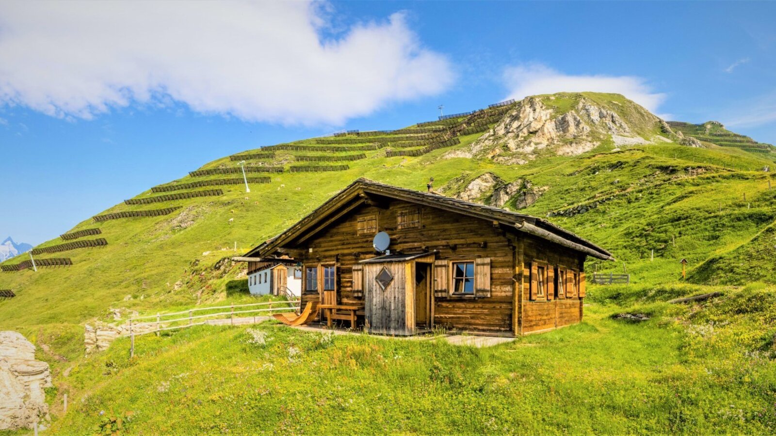 Die Almhütte in Holzbauweise liegt auf einem grünen Berghang und bietet einen Sitzbereich im Freien.