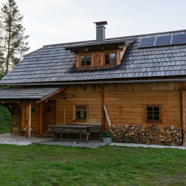Die Alm in Holzbauweise mit Schindeldach, Solarpaneelen und einem überdachten Sitzbereich mit Picknickbank und gestapeltem Brennholz.