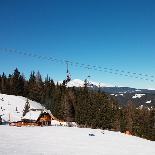 Skigebiet mit Skipiste, Skilift und einer Almhütte vor verschneiter Berglandschaft.