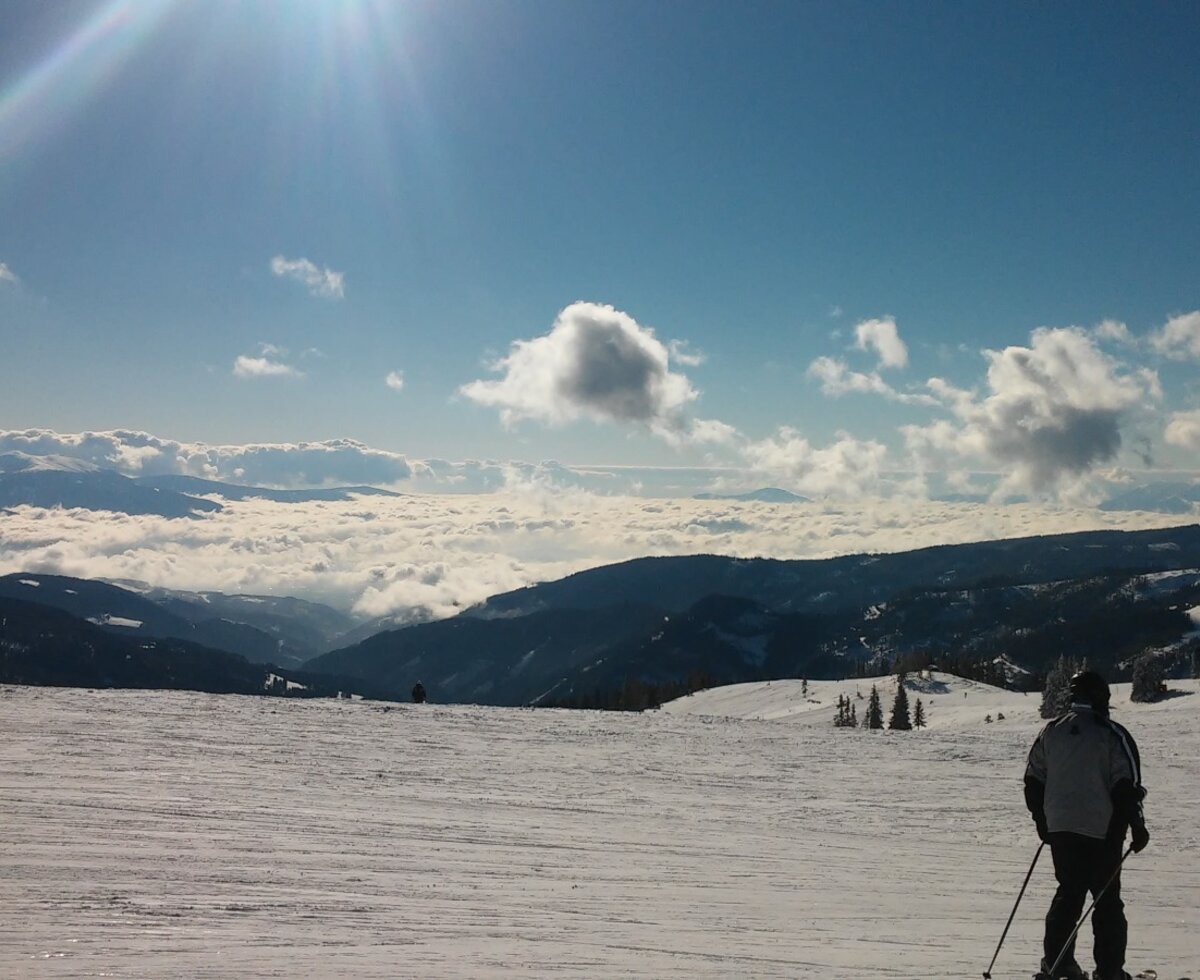 Winterliche Landschaft rund um die Alm mit Skifahrern auf der Piste und Panoramablick über eine Wolkendecke.