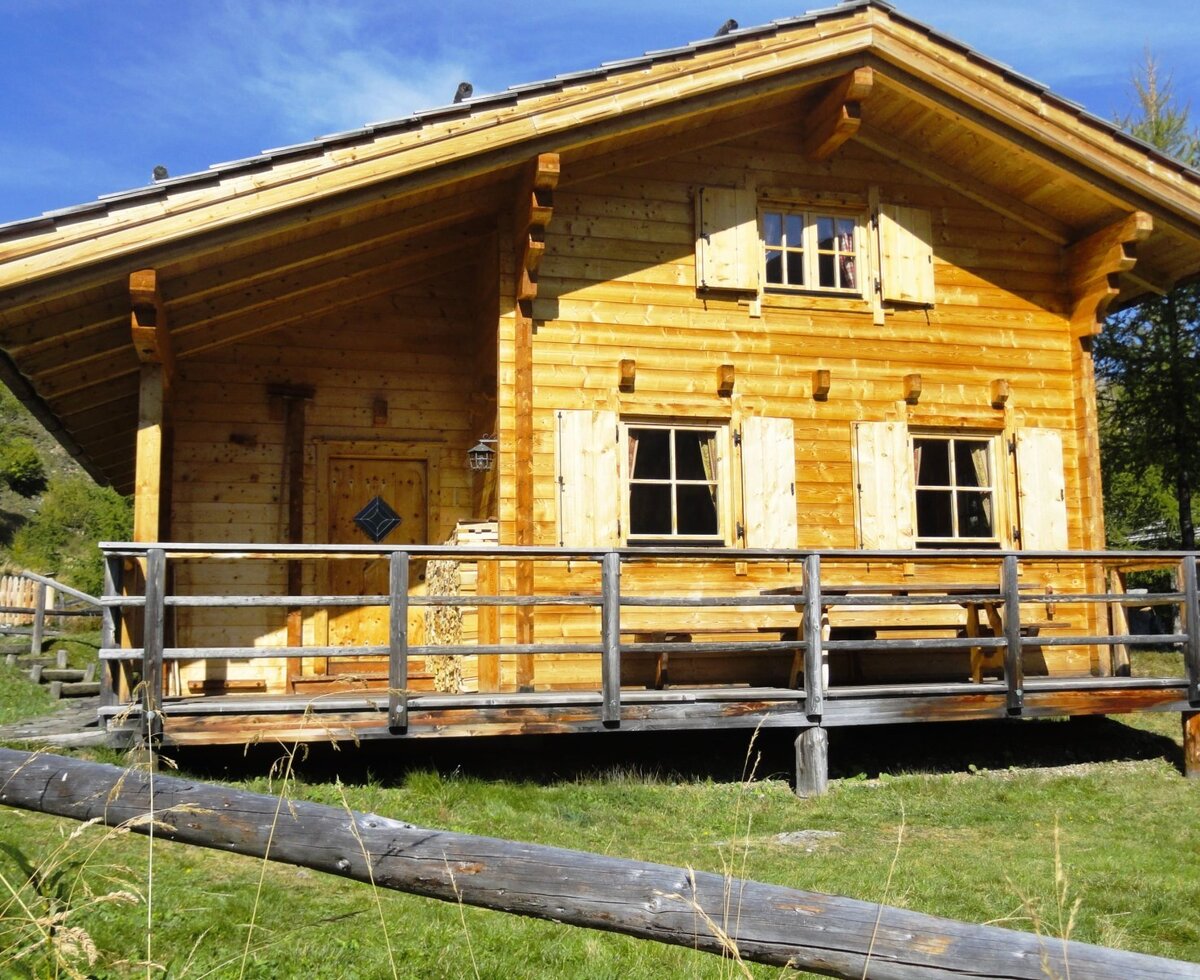 Die Alm mit Holzfassade, einem erhöhten Balkon inklusive Sitzgelegenheit und Fenstern mit Holzläden.