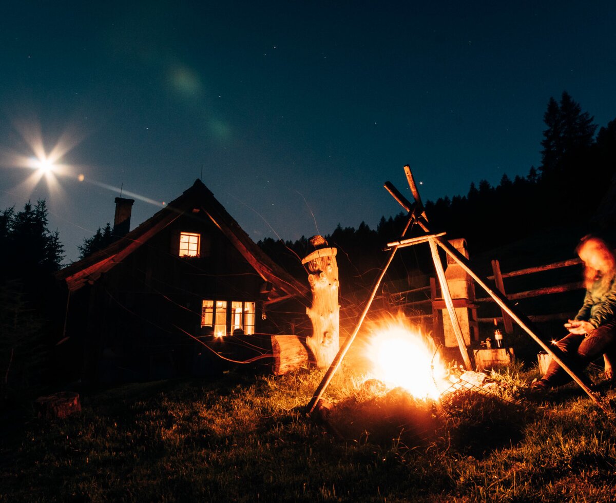 Lagerfeuer vor der Alm bei Nacht, mit beleuchteten Fenstern und dem Mond am Himmel.