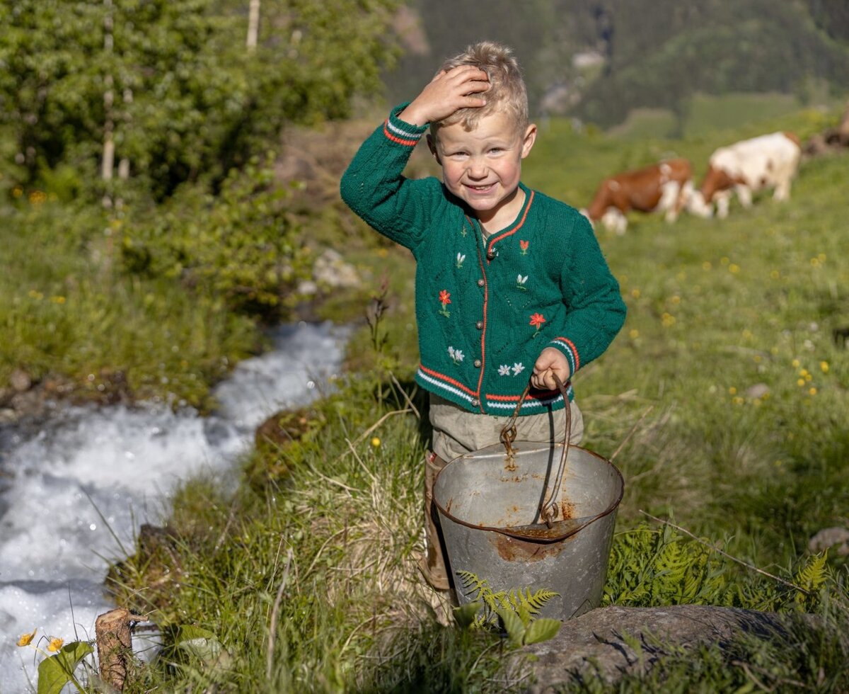 Ein Kind steht mit einem Eimer an einem Gebirgsbach auf der Alm, mit grasenden Kühen im Hintergrund.