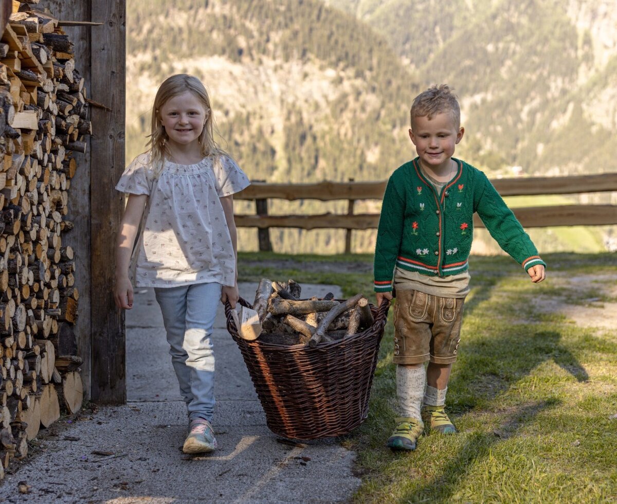 Kinder tragen Brennholz auf der Alm, mit einem Holzstapel und Bergblick.