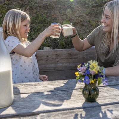 Zwei Personen stoßen mit frischer Milch an einem Holztisch auf der Alm an.