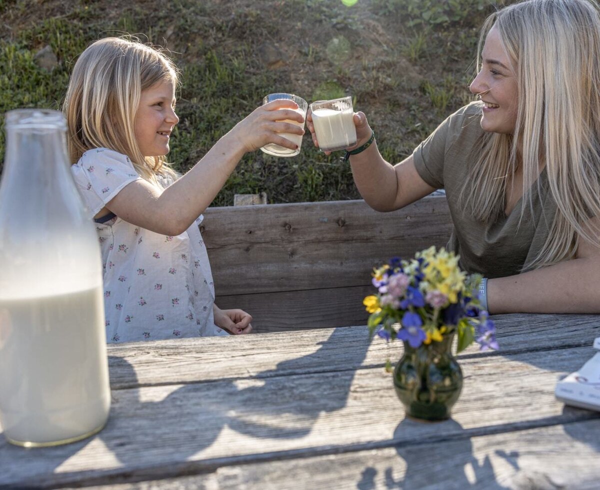 Zwei Personen stoßen mit frischer Milch an einem Holztisch auf der Alm an.