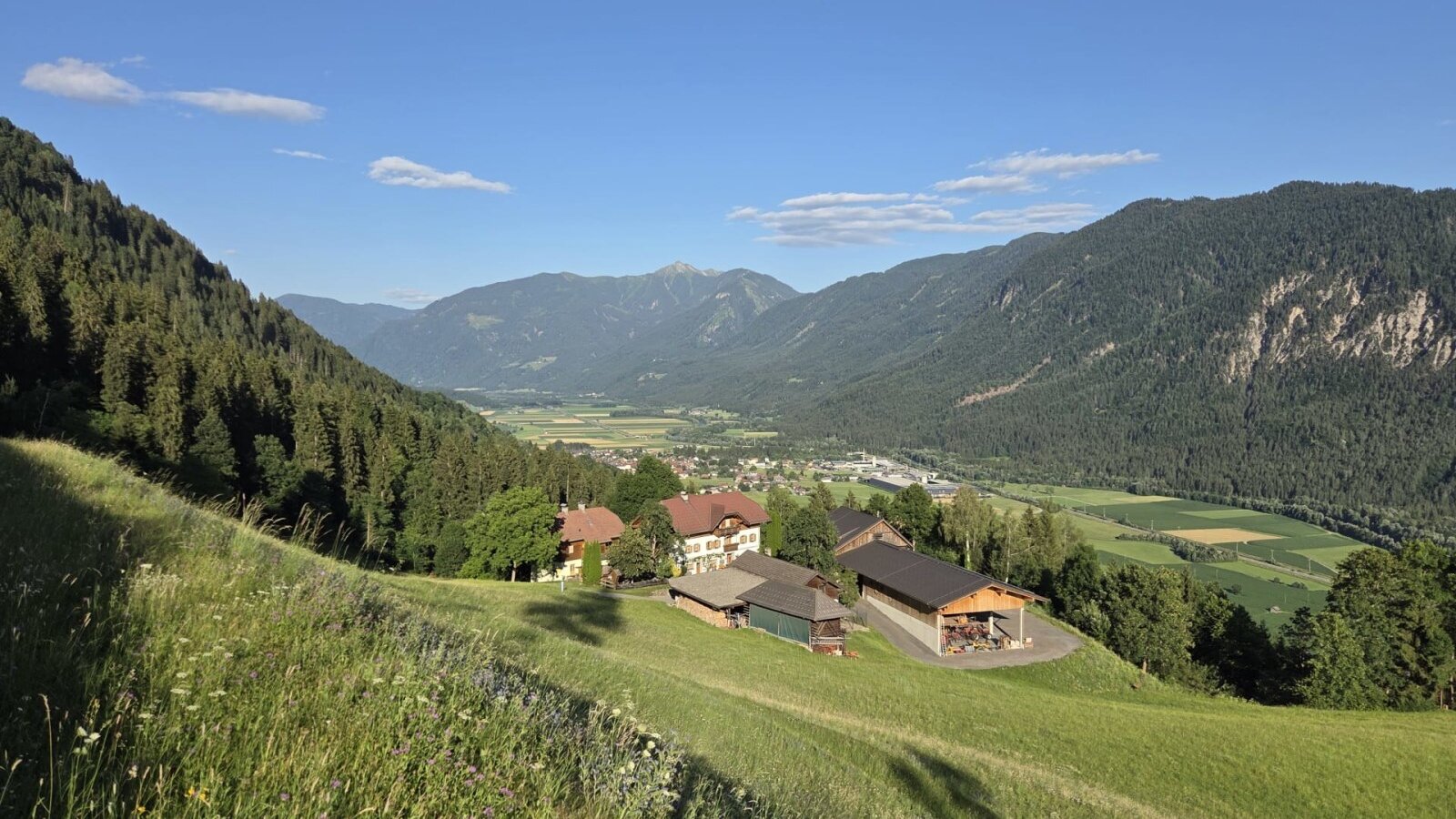 Die idyllische Lage des Bauernhofs an einem saftig grünen Hang mit weitem Ausblick auf das Tal und die umliegenden Berge.