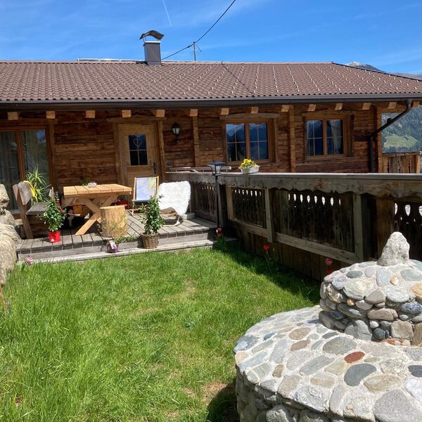 Die Alm mit einer Holzterrasse, ausgestattet mit Tisch und Stühlen, umgeben von einer grünen Wiese und Bergblick.