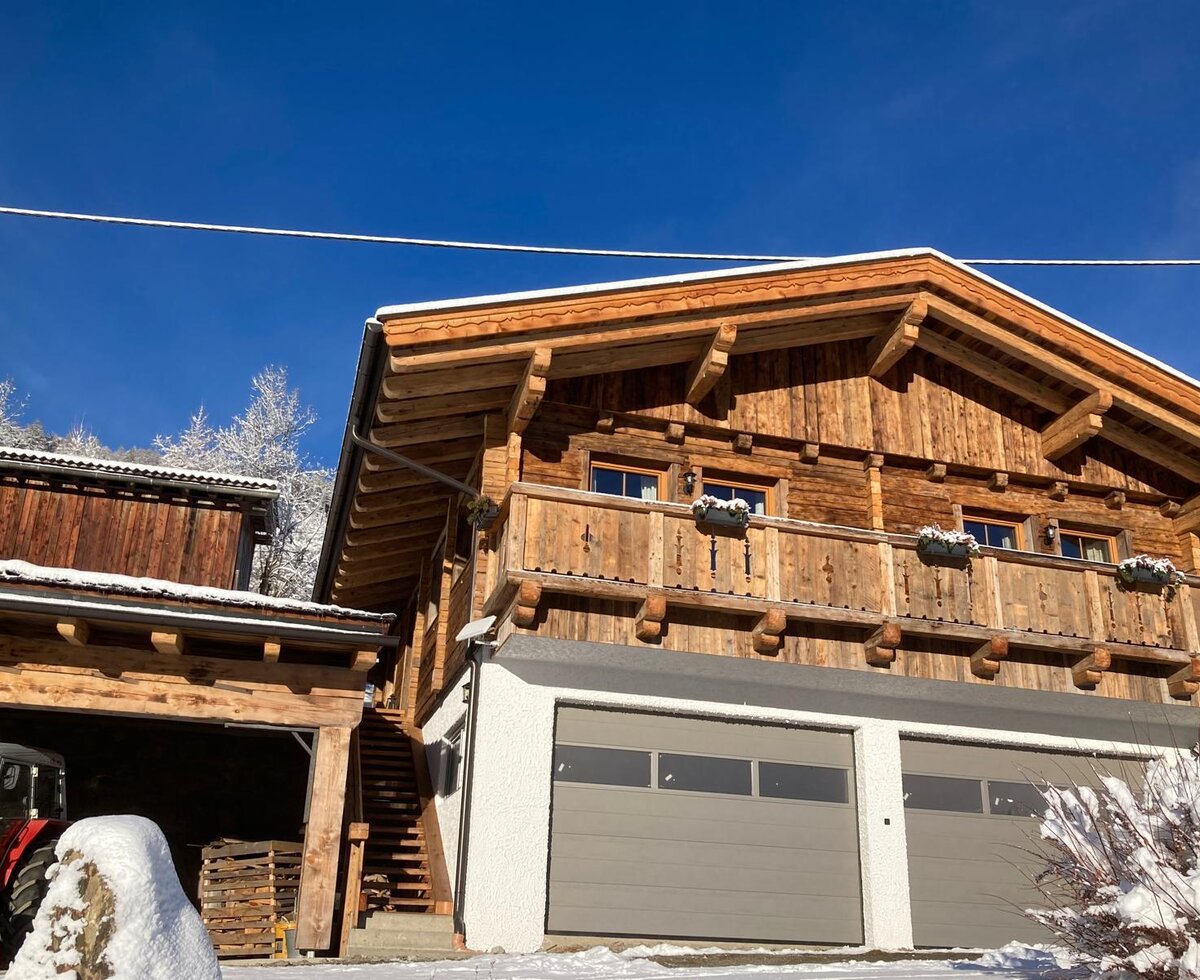 Außenansicht der Alm mit Holzbalkon, Fenstern, zwei Garagentoren und einer Außentreppe, umgeben von Schnee.