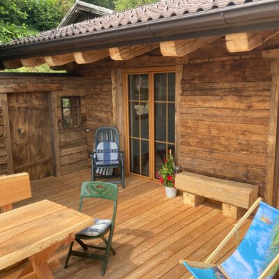 Die Holzterrasse der Alm mit Esstisch, verschiedenen Sitzgelegenheiten und Liegestuhl.