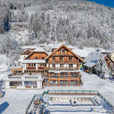 Außenansicht des Bauernhofs im Winter, umgeben von Schnee und Bergen, mit Balkonen und einem Kinderspielbereich.