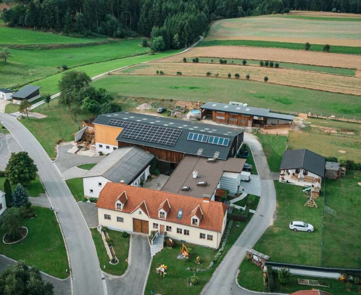 Luftaufnahme des Alpakahof-Anwesens mit mehreren Gebäuden, Solaranlagen auf den Dächern und umgeben von Feldern und Wäldern.
