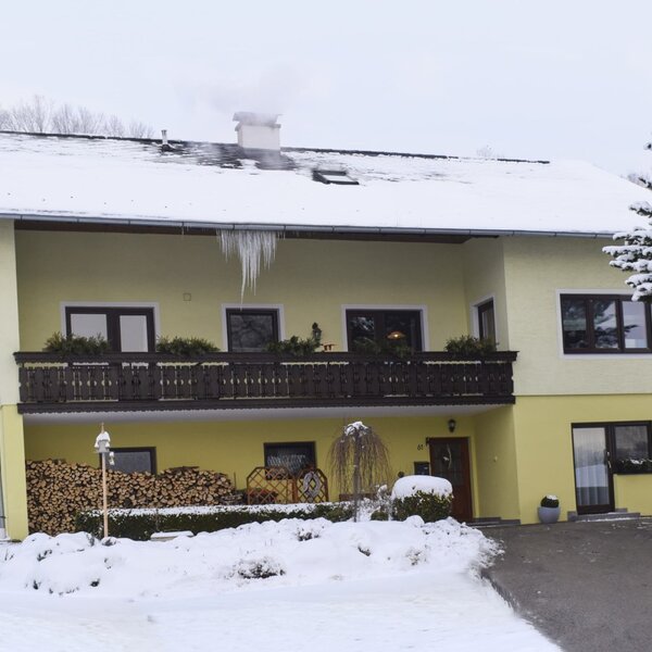 Der winterliche Bauernhof von außen, mit Schnee auf dem Dach und Boden, einem rauchenden Schornstein, Balkon und Holzstapel.