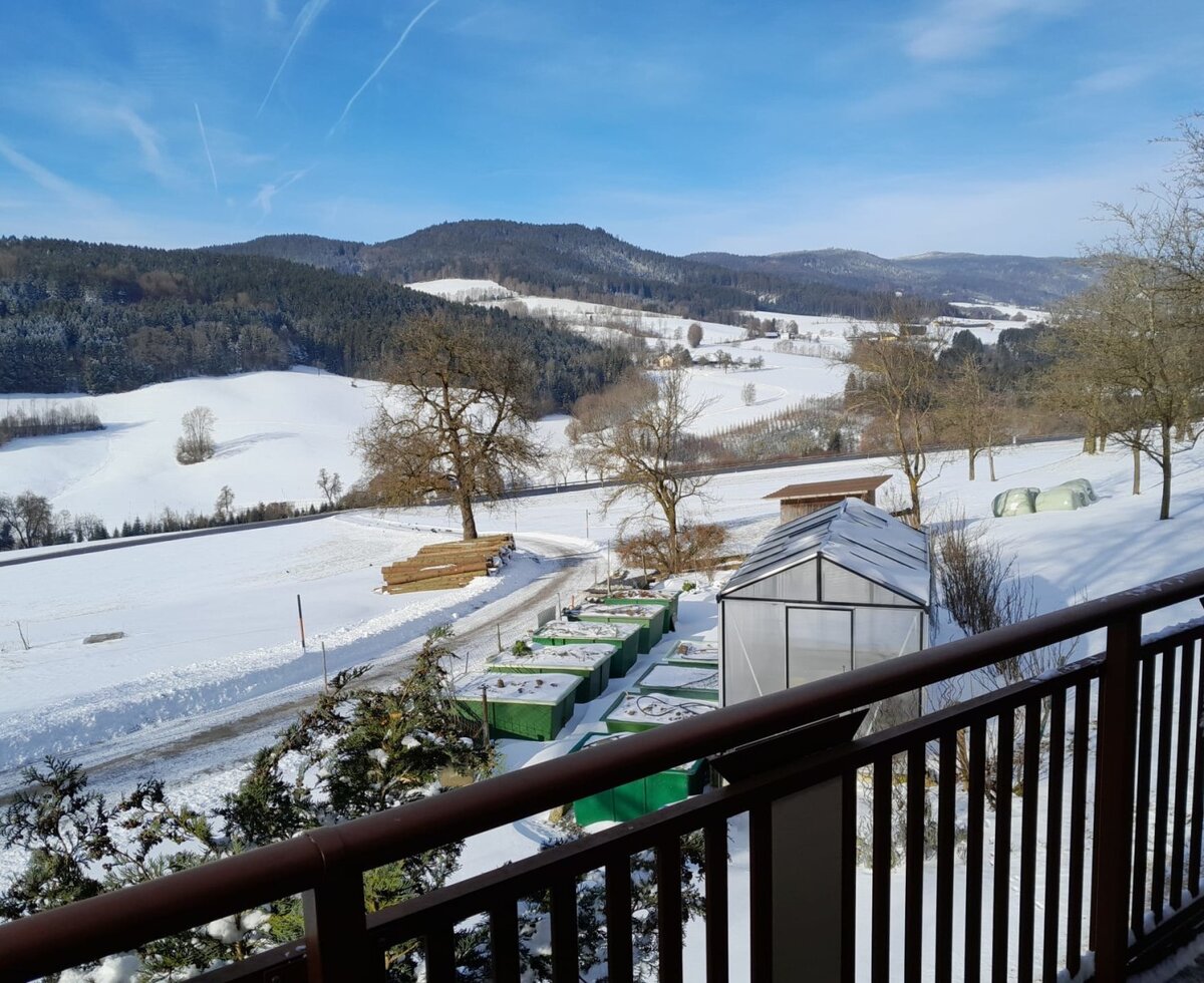 Blick vom Balkon des Bauernhofs auf die verschneite Winterlandschaft mit Hügeln, Wäldern, einem Gewächshaus und landwirtschaftlichen Behältern.