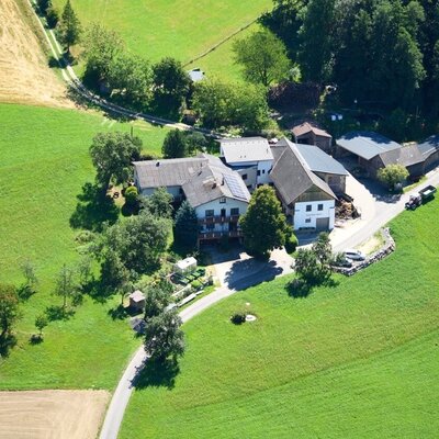 Flugaufnahme vom Bauernhof Waira mit seinen Gebäuden, umgeben von grünen Wiesen und Bäumen, mit einer Zufahrtsstraße.