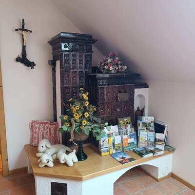 Traditioneller Kachelofen mit Sitzbank, dekoriert mit Blumen, Stofftieren und Informationsmaterial für Gäste auf dem Bauernhof.
