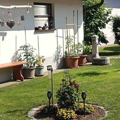 Gartenbereich des Bauernhofs mit Rasenfläche, Blumenbeet, einer Holzbank, Topfpflanzen und einem Steinbrunnen.