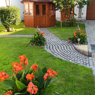 Gartenbereich des Bauernhofs mit einer Holzgartenlaube, einem gepflasterten Weg und blühenden Pflanzen.
