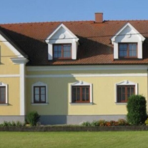 Die Außenansicht des Bauernhofs mit gelber Fassade, braunem Ziegeldach, Fenstern und einer großen Holztür.