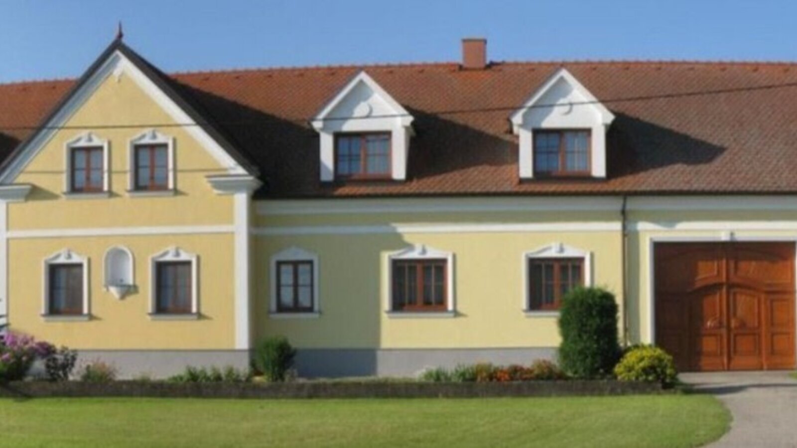 Die Außenansicht des Bauernhofs mit gelber Fassade, braunem Ziegeldach, Fenstern und einer großen Holztür.
