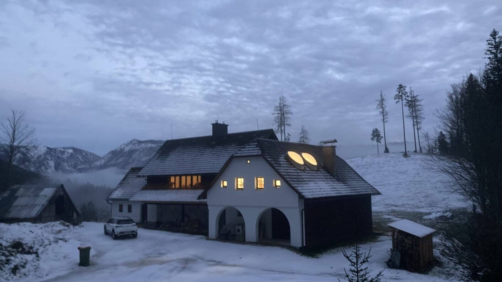 Außenansicht im Winter mit Schnee, beleuchteten Fenstern und Bergblick.