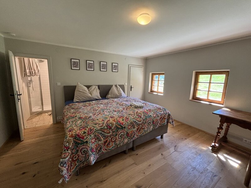 Doppelzimmer mit Holzboden, zwei Fenstern und Zugang zu einem Badezimmer mit Dusche.