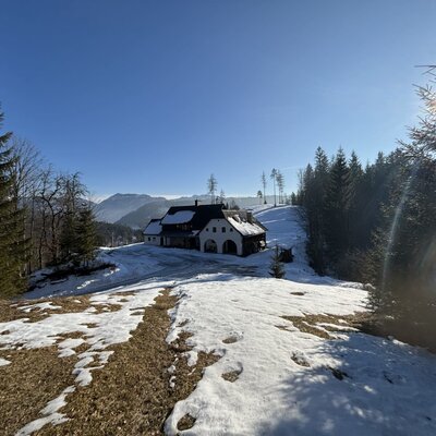 Die Almhütte in einer verschneiten Berglandschaft mit Wald unter blauem Himmel.