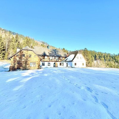 Außenansicht in einer verschneiten Winterlandschaft mit angrenzendem Wald und Bergen im Hintergrund.