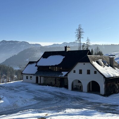 Die Almhütte im Winter mit schneebedecktem Dach und einer Berglandschaft im Hintergrund.