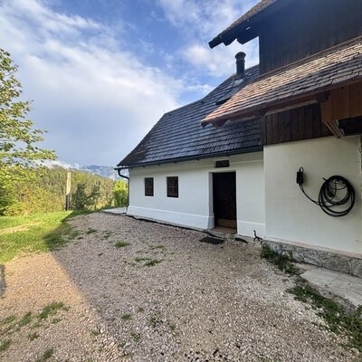 Außenansicht mit weißer Fassade, Holzdach und Schotterweg, umgeben von Bäumen und Bergen.