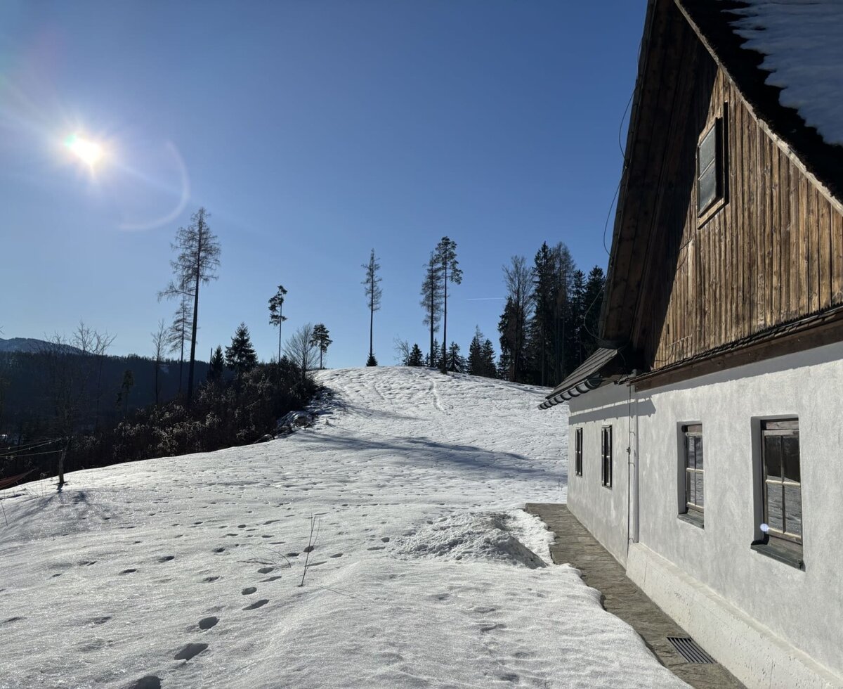 Die Almhütte im Winter mit schneebedecktem Dach und weißer Fassade, umgeben von einer verschneiten Hügellandschaft und Bäumen.
