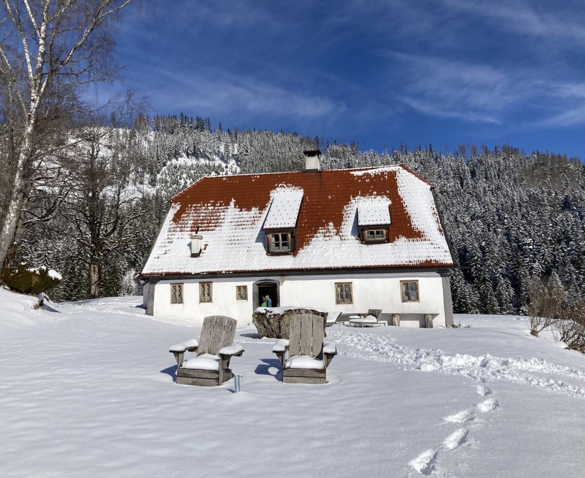 Das Bed and Breakfast mit schneebedecktem roten Dach und weißer Fassade, umgeben von einem verschneiten Wald und Bergen, mit zwei Holzstühlen im Schnee.