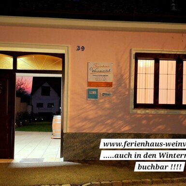 Die beleuchtete Fassade des Bauernhofs bei Nacht mit offenem Eingang, festlichen Dekorationen und Blick in den Garten.