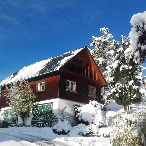 Der Bauernhof im Winter, umgeben von verschneiten Bäumen und einer kleinen Holzlaube auf einem Hügel unter blauem Himmel.