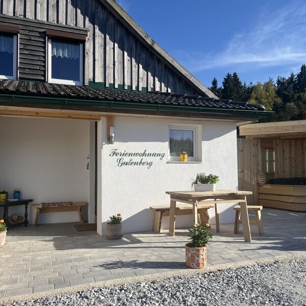 Außenansicht des Bauernhofs mit dem Eingang zur Ferienwohnung Gutenberg, einer Terrasse mit Holztisch und Bänken sowie einem überdachten Außen-Whirlpool.