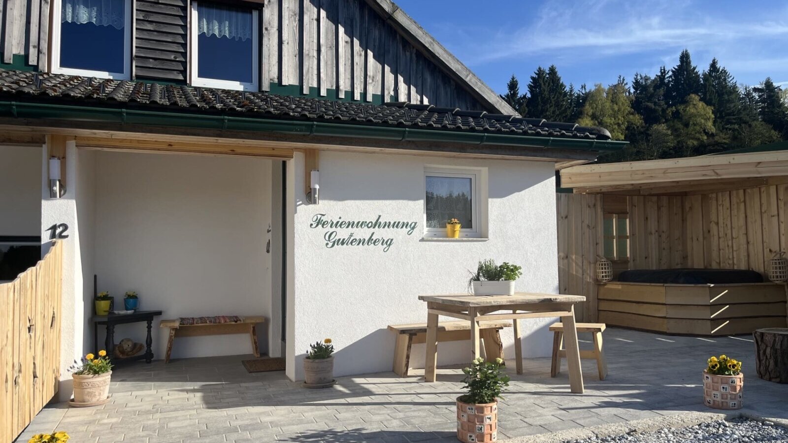 Außenansicht des Bauernhofs mit dem Eingang zur Ferienwohnung Gutenberg, einer Terrasse mit Holztisch und Bänken sowie einem überdachten Außen-Whirlpool.