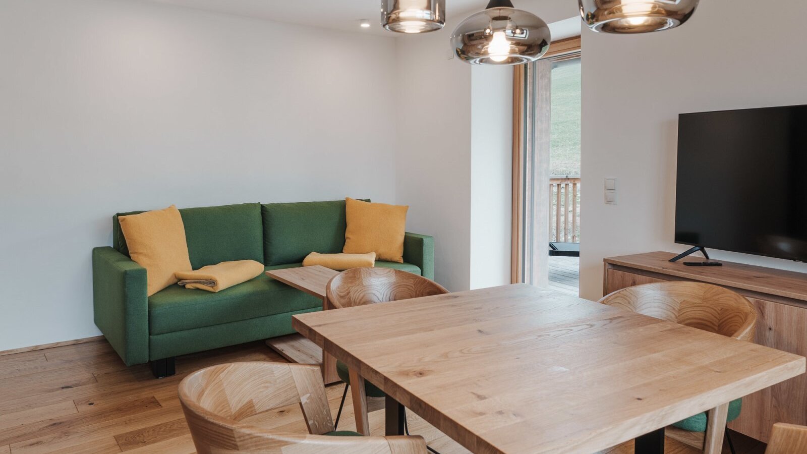 Einladender Wohnbereich des Landhofs mit einem massiven Holztisch, gemütlichem grünem Sofa und modernem Fernseher.