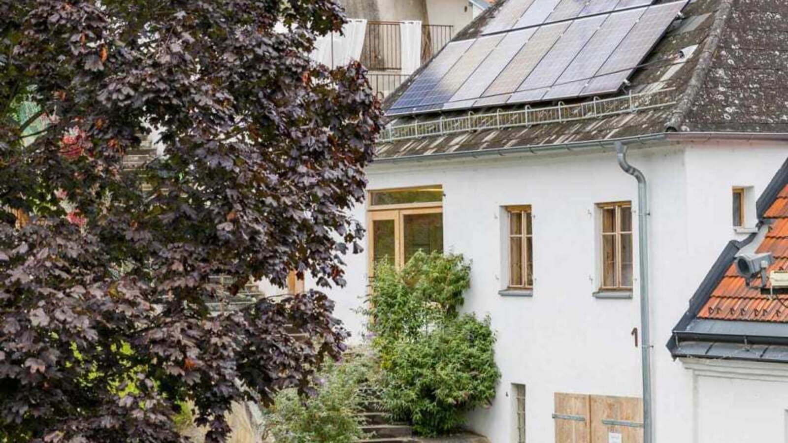 Die Außenansicht der Greisslerei mit weißer Fassade, Solarpaneelen auf dem Dach, einer Holzpforte und Außentreppe.
